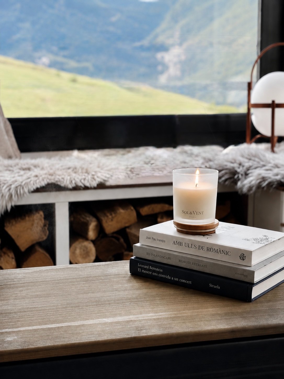 Vela Sol&Vent encendida sobre unos libros junto a una ventana con vistas a la montaña
